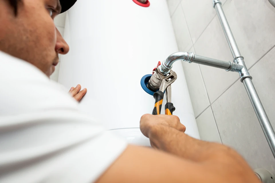 Photographie professionnelle d'un technicien plombier en tenue de travail inspectant ou manipulant un chauffe-eau mural dans une salle de bain moderne. Le chauffe-eau doit être clairement visible au centre de l'image. La scène capture le professionnel en action, concentré sur son travail, dans un environnement résidentiel réaliste et bien éclairé.